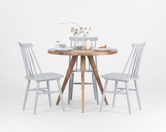 May include: A round wooden table with a gray top and a gray painted base. Four gray chairs surround the table. The table is set with plates, glasses, and a vase with dried branches.