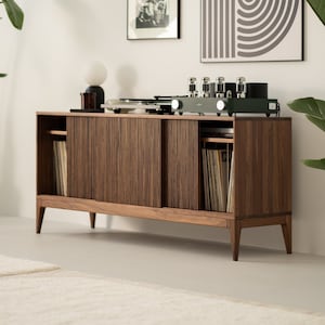 May include: A mid-century modern style wooden record console with a turntable, amplifier, and sliding doors. The console is filled with vinyl records and is set against a white wall with artwork and plants.
