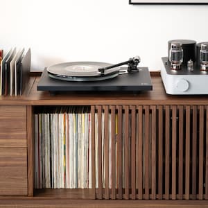 May include: A black turntable with a record on it, sitting on a wooden cabinet with a record collection and a white stereo amplifier. The cabinet has a drawer and a shelf with books. A poster with the word "bauhaus" is on the wall behind the turntable.