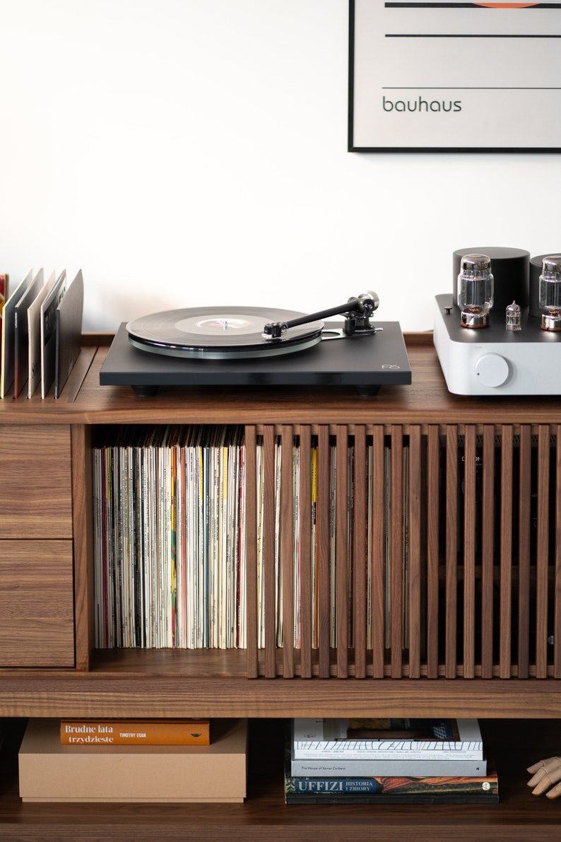 May include: A black turntable with a record on it, sitting on a wooden cabinet with a record collection and a white stereo amplifier. The cabinet has a drawer and a shelf with books. A poster with the word "bauhaus" is on the wall behind the turntable.