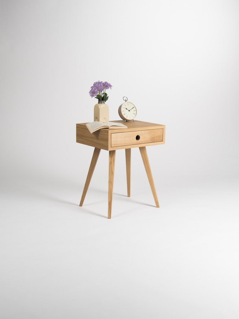 May include: A light wood bedside table with a single drawer and four legs. A small clock and a vase with purple flowers are on top of the table.