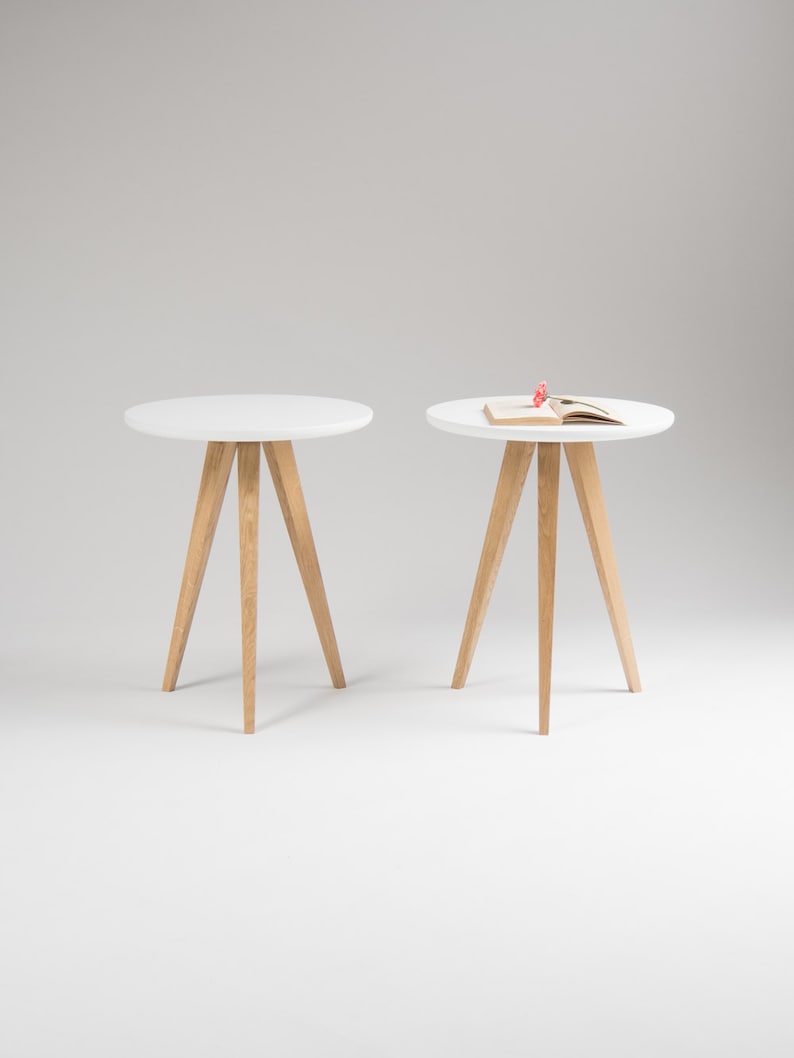 May include: Two white round side tables with wooden legs. The table on the right has a book on top.