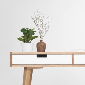 Puede incluir: Un escritorio blanco con dos cajones y patas de madera. Una peque&ntilde;a planta en maceta y un jarr&oacute;n con ramas secas est&aacute;n encima del escritorio.