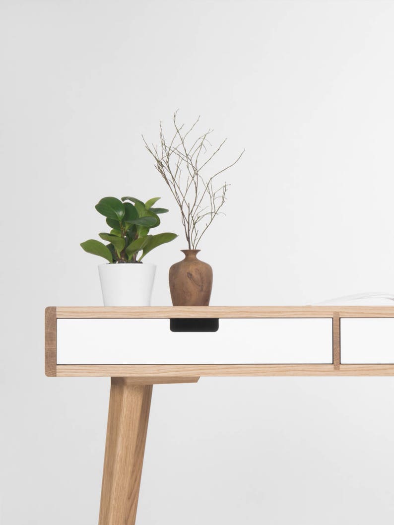 Puede incluir: Un escritorio blanco con dos cajones y patas de madera. Una peque&ntilde;a planta en maceta y un jarr&oacute;n con ramas secas est&aacute;n encima del escritorio.