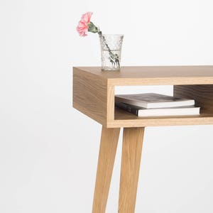 May include: A light wood side table with a shelf and four legs. A pink carnation in a clear glass vase sits on the top shelf.