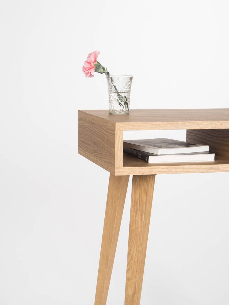 May include: A light wood side table with a shelf and four legs. A pink carnation in a clear glass vase sits on the top shelf.