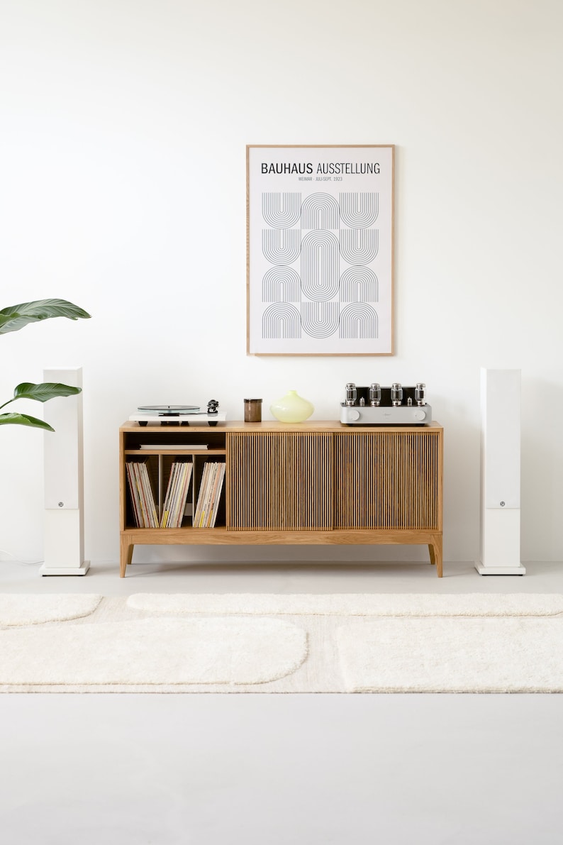 Puede incluir: Un armario de madera con dos puertas y un estante para discos de vinilo. Un tocadiscos, un amplificador y altavoces est&aacute;n encima del armario. Un p&oacute;ster enmarcado con el texto "BAUHAUS AUSSTELLUNG" est&aacute; en la pared detr&aacute;s del armario. Una alfombra blanca est&aacute; en el suelo delante del armario.