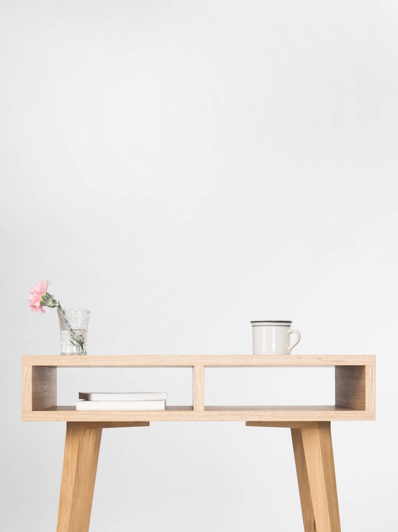 May include: A light wood console table with two open shelves and tapered legs. A pink carnation in a glass vase and a white mug are on the top shelf.
