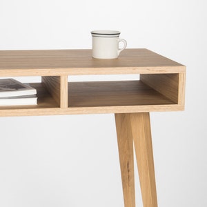 May include: A light wood desk with a shelf and three legs. A white mug with a black stripe sits on the desk.