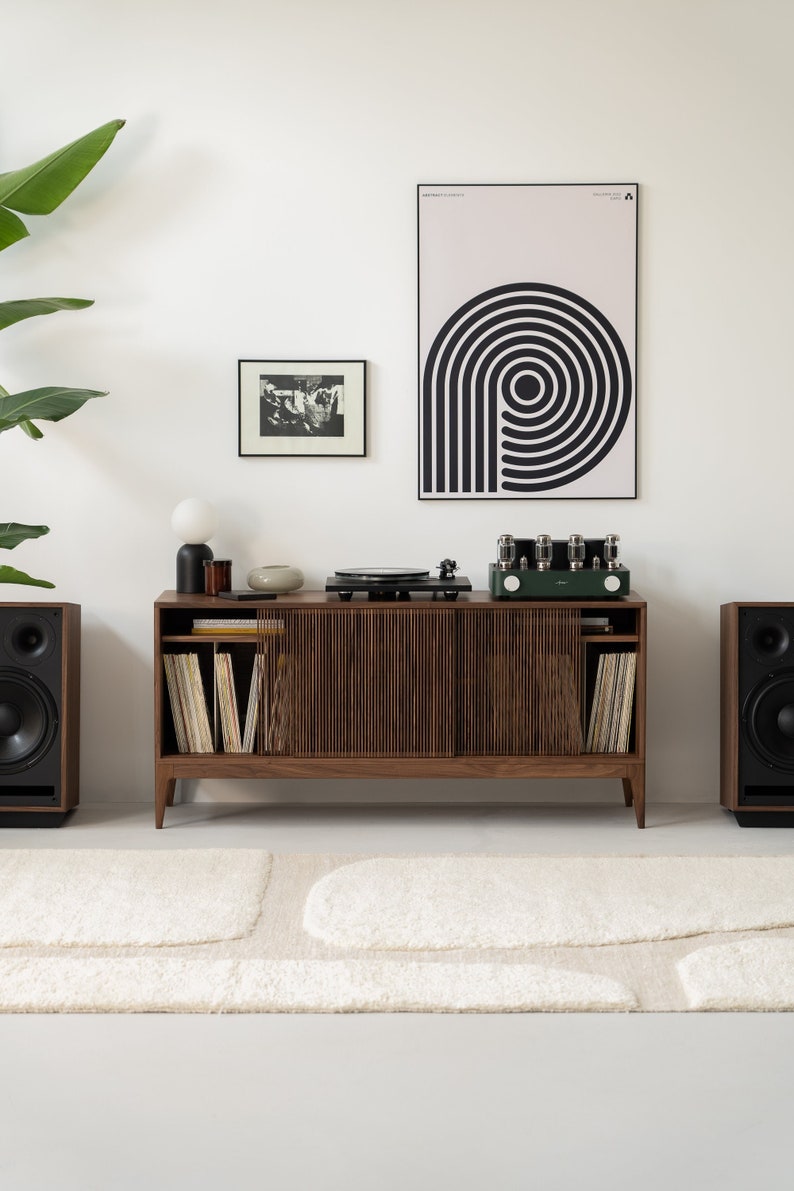 May include: A mid-century modern style entertainment center with a record player, speakers, and a green amplifier. The wooden cabinet has a slatted front and holds vinyl records. A black and white art print hangs on the wall.