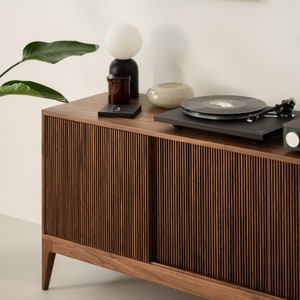 May include: A mid-century modern style wooden cabinet with a record player on top. The cabinet has a vertical slat design on the sliding doors. A white globe lamp, a small glass vase, and a framed black and white print are also visible.