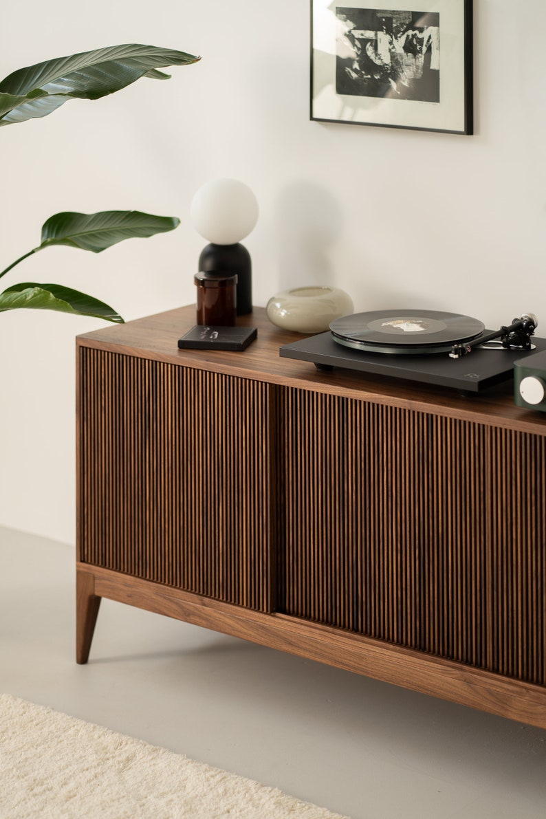 May include: A mid-century modern style wooden cabinet with a record player on top. The cabinet has a vertical slat design on the sliding doors. A white globe lamp, a small glass vase, and a framed black and white print are also visible.