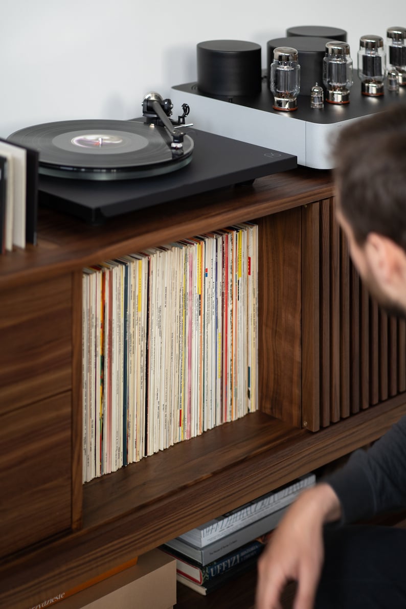 May include: A wooden cabinet with a record player on top and a shelf full of vinyl records. The cabinet has a drawer on the left side and a door on the right side. The door is open and the shelves are full of records. The record player is black and has a silver tone arm. There is a stack of books on the bottom shelf.