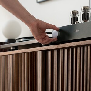 May include: A hand adjusting a silver knob on a black audio amplifier. The amplifier sits atop a wooden cabinet with vertical slat detailing. Two glass tubes are visible on the amplifier. A turntable and a white globe lamp are also in the image.