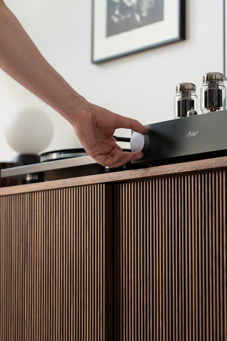 May include: A hand adjusting a silver knob on a black audio amplifier. The amplifier sits atop a wooden cabinet with vertical slat detailing. Two glass tubes are visible on the amplifier. A turntable and a white globe lamp are also in the image.