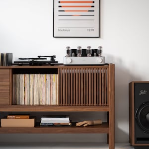 May include: A wooden cabinet with a record player, amplifier, and speaker. The cabinet has a shelf with books and a wooden hand. The cabinet is in front of a white wall with a framed print that says "bauhaus 1919".