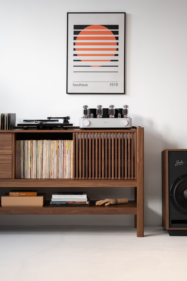 May include: A wooden cabinet with a record player, amplifier, and speaker. The cabinet has a shelf with books and a wooden hand. The cabinet is in front of a white wall with a framed print that says "bauhaus 1919".