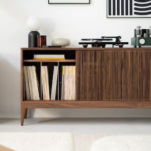 May include: A mid-century modern style wooden record cabinet with a built-in record player. The cabinet has a slatted door and open shelving for vinyl records. A black and white framed art piece hangs on the wall.