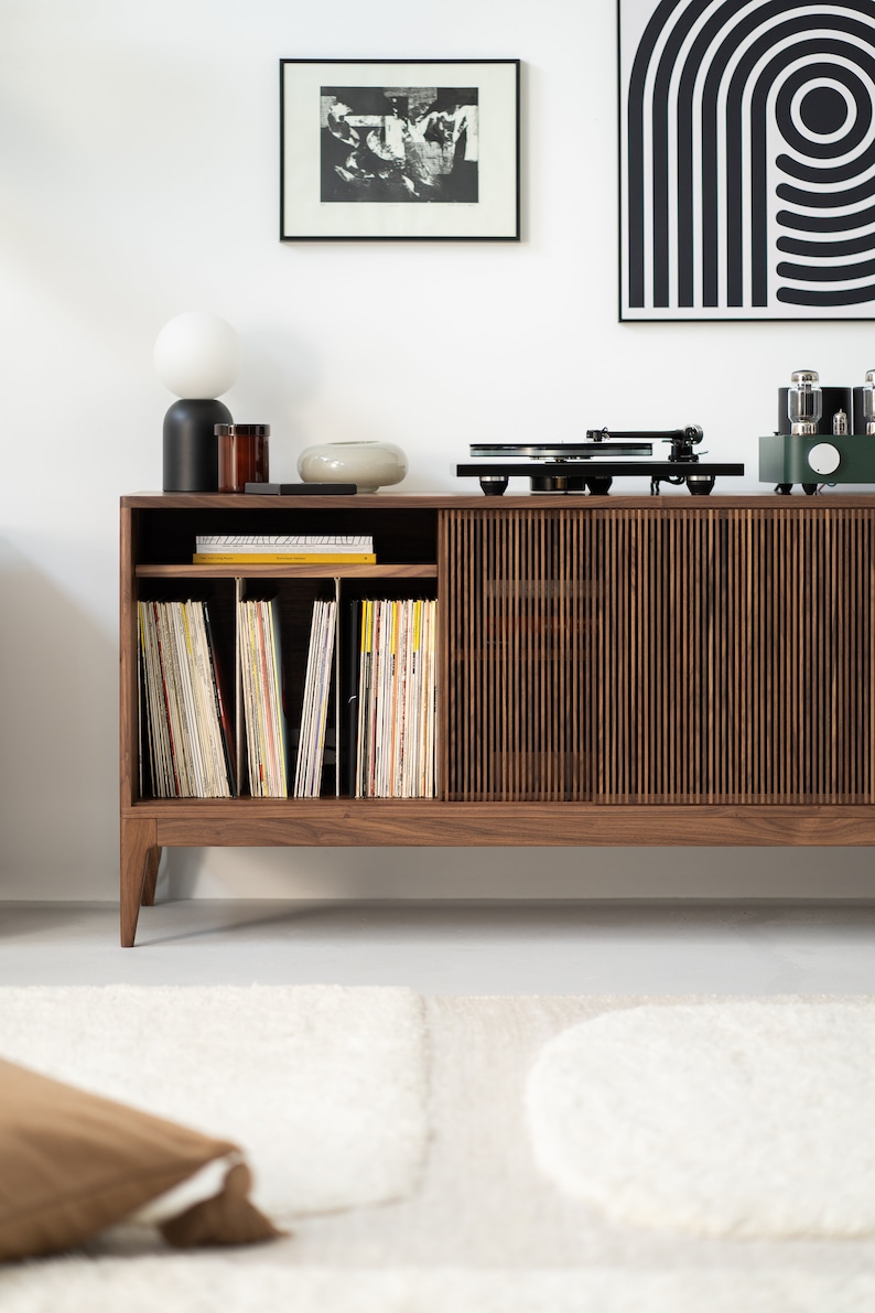 May include: A mid-century modern style wooden record cabinet with a built-in record player. The cabinet has a slatted door and open shelving for vinyl records. A black and white framed art piece hangs on the wall.