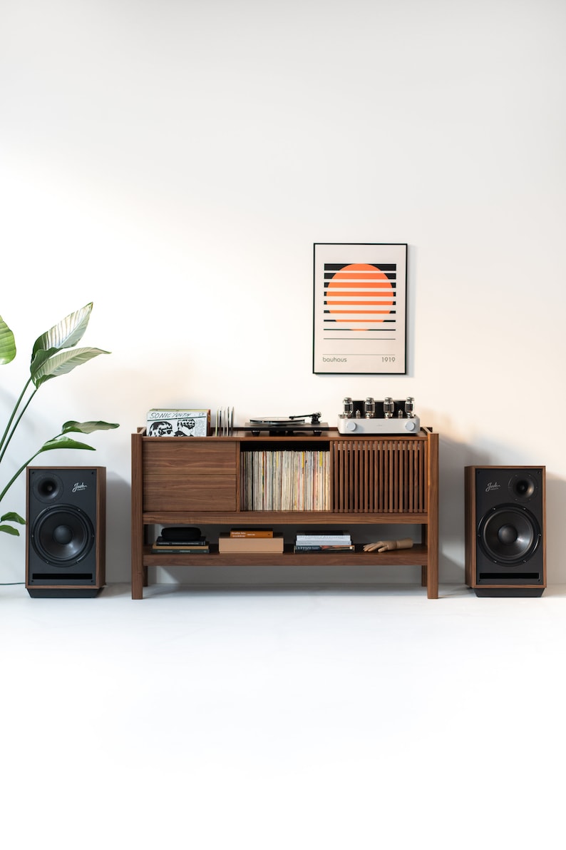May include: A wooden record player stand with a turntable, amplifier, and speakers. The stand has a shelf for records and a drawer for storage. There is a framed print with the text "bauhaus 1919" above the stand.