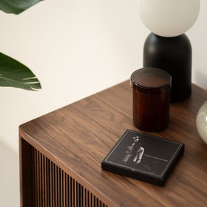 May include: A modern, minimalist scene featuring a dark wood table with a slatted side. On the table are a black and white photograph, a brown jar with a lid, and a black and white lamp.