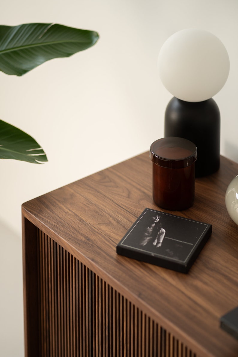 May include: A modern, minimalist scene featuring a dark wood table with a slatted side. On the table are a black and white photograph, a brown jar with a lid, and a black and white lamp.