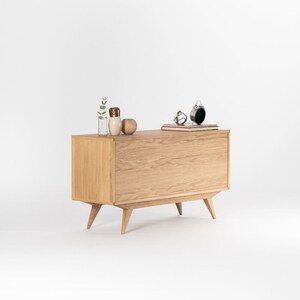 May include: A light wood cabinet with two doors and four legs. The cabinet has a modern design with a slightly angled front and a simple, clean look. The cabinet is sitting on a white surface.