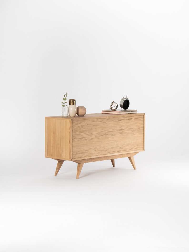 May include: A light wood cabinet with two doors and four legs. The cabinet has a modern design with a slightly angled front and a simple, clean look. The cabinet is sitting on a white surface.