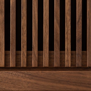 May include: Close-up of a wooden panel featuring vertical slats in a warm brown tone. The slats are evenly spaced, creating a rhythmic pattern against a dark background. The wood grain is visible, adding texture and depth to the design. The panel has a horizontal base.