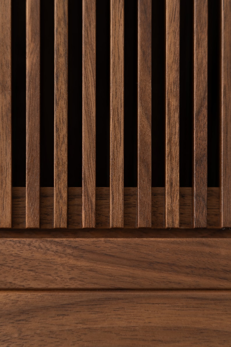 May include: Close-up of a wooden panel featuring vertical slats in a warm brown tone. The slats are evenly spaced, creating a rhythmic pattern against a dark background. The wood grain is visible, adding texture and depth to the design. The panel has a horizontal base.