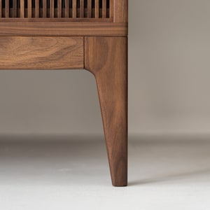 May include: Close-up of a wooden cabinet with a slatted front and a single drawer. The cabinet is made of dark brown wood and has a tapered leg. The background is a neutral color.