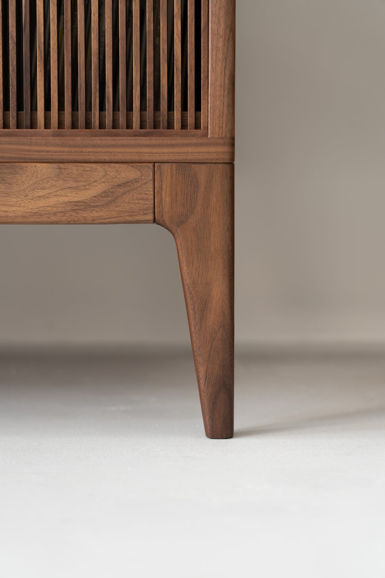 May include: Close-up of a wooden cabinet with a slatted front and a single drawer. The cabinet is made of dark brown wood and has a tapered leg. The background is a neutral color.