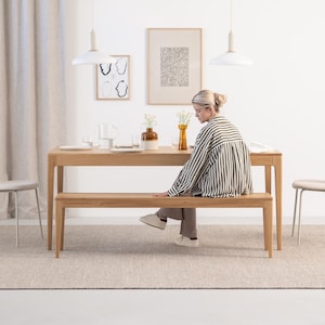 May include: A light wood dining table and bench set with a person sitting on the bench. The table and bench are both made of natural wood and have a simple, modern design. The table is rectangular and has a light wood finish. The bench is also rectangular and has a light wood finish. The person is wearing a striped shirt and is sitting on the bench with their legs crossed. The table and bench are set in a room with white walls and a light brown rug.