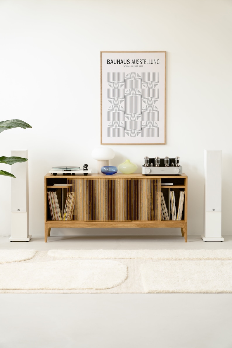 Puede incluir: Un armario de madera de estilo moderno de mediados de siglo con puertas correderas, que incluye un tocadiscos, un amplificador vintage y dos altavoces blancos. El armario est&aacute; decorado con un p&oacute;ster enmarcado que dice "BAUHAUS AUSSTELLUNG WEIMAR, 20. AUG. - SEPT. 1923".