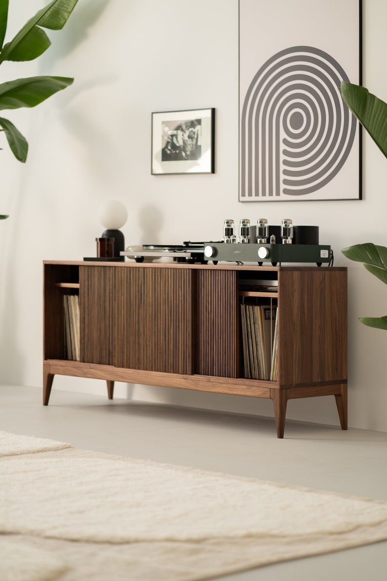 May include: A mid-century modern style wooden record console with a turntable, amplifier, and sliding doors. The console is filled with vinyl records and is set against a white wall with artwork and plants.