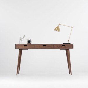 Puede incluir: Un escritorio de madera con tres cajones y una l&aacute;mpara de escritorio dorada. El escritorio tiene un dise&ntilde;o minimalista con patas c&oacute;nicas y un acabado en madera oscura.