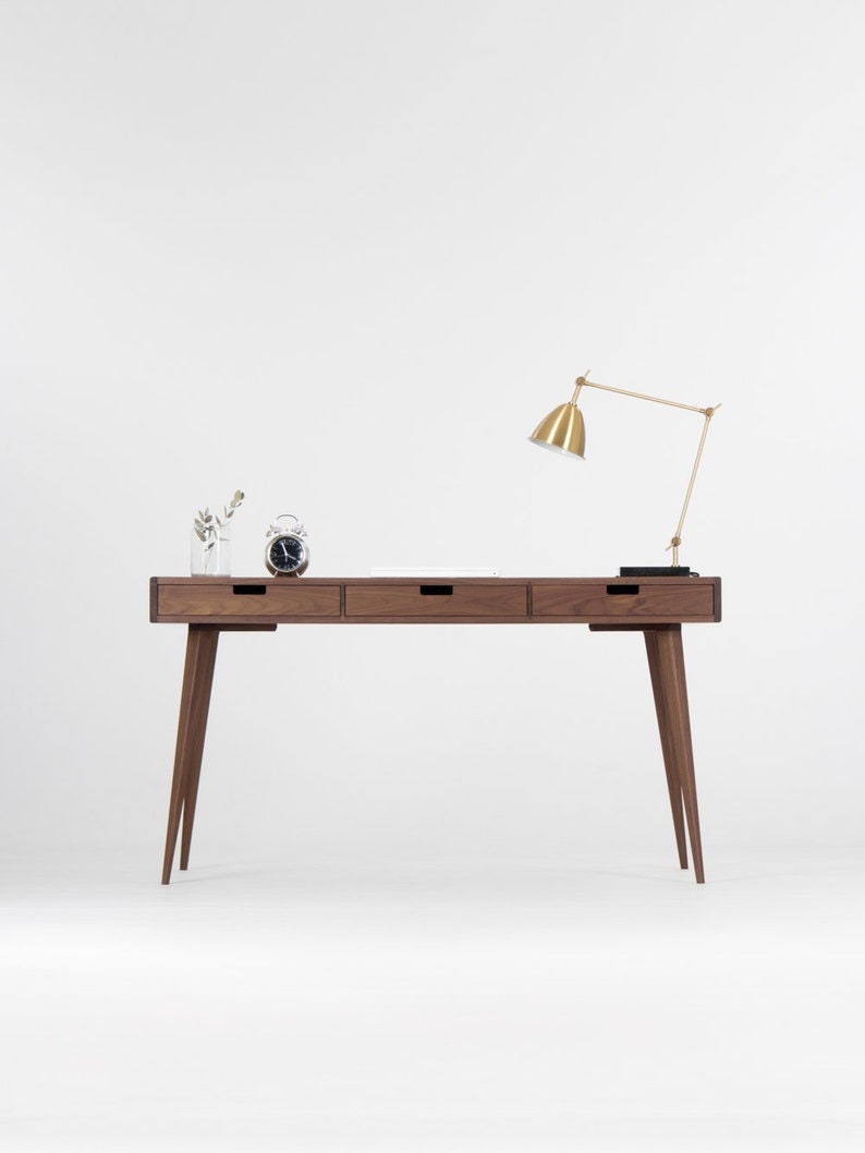 Puede incluir: Un escritorio de madera con tres cajones y una l&aacute;mpara de escritorio dorada. El escritorio tiene un dise&ntilde;o minimalista con patas c&oacute;nicas y un acabado en madera oscura.