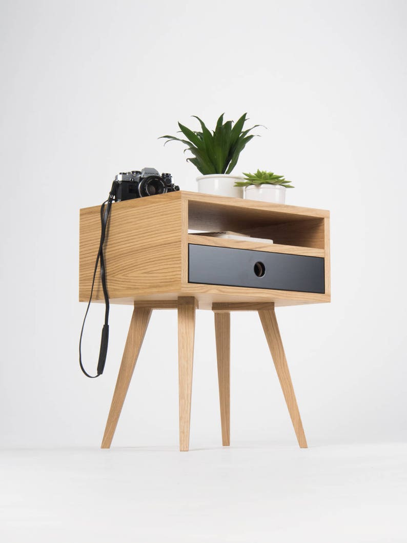 Puede incluir: Una mesita de noche de madera con un caj&oacute;n negro y un estante. La mesita de noche tiene cuatro patas y est&aacute; decorada con una c&aacute;mara y dos plantas en macetas.