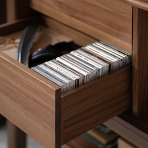 May include: A wooden cabinet with a drawer open, revealing a stack of CDs. The drawer is made of dark wood and has a smooth finish. The CDs are arranged neatly in the drawer, with their spines facing out.