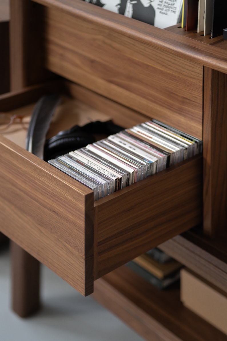 May include: A wooden cabinet with a drawer open, revealing a stack of CDs. The drawer is made of dark wood and has a smooth finish. The CDs are arranged neatly in the drawer, with their spines facing out.