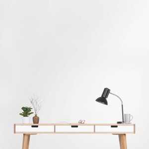 Puede incluir: Un escritorio blanco con tres cajones y patas de madera. Una l&aacute;mpara de escritorio negra con una pantalla blanca est&aacute; sobre el escritorio. Hay dos peque&ntilde;as plantas en maceta y un par de gafas sobre el escritorio.