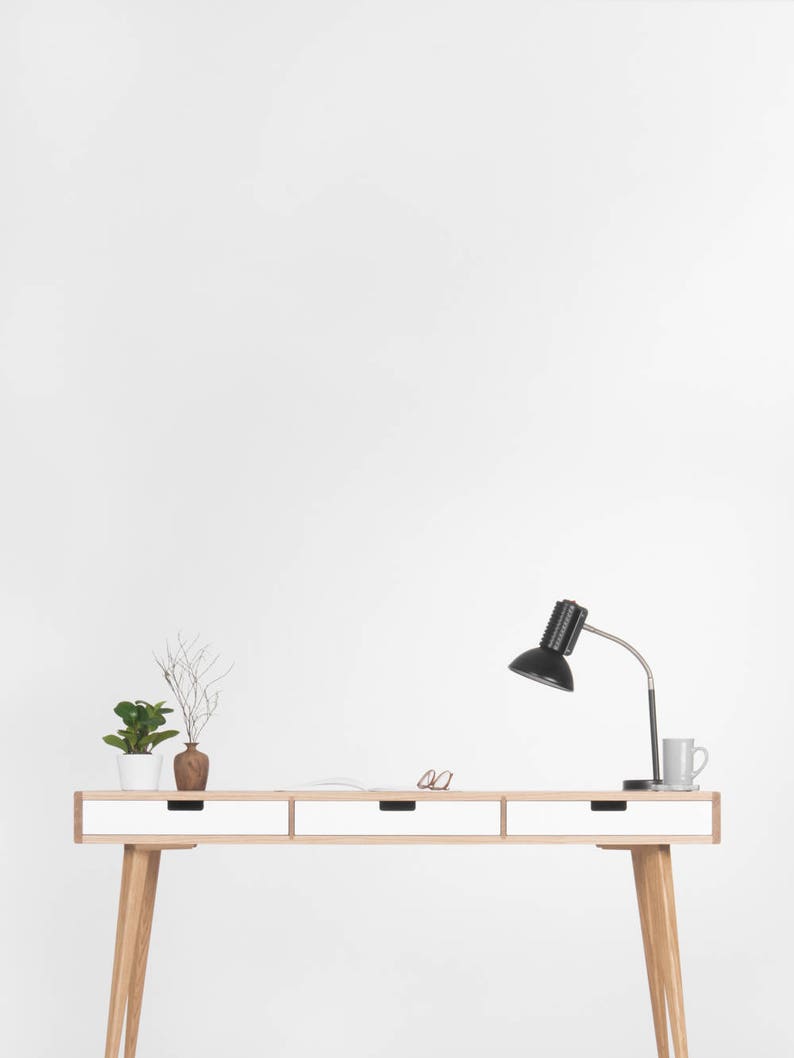 Puede incluir: Un escritorio blanco con tres cajones y patas de madera. Una l&aacute;mpara de escritorio negra con una pantalla blanca est&aacute; sobre el escritorio. Hay dos peque&ntilde;as plantas en maceta y un par de gafas sobre el escritorio.