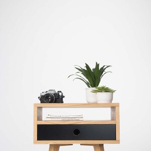 Puede incluir: Una mesita de noche de madera con un caj&oacute;n negro y un estante. La mesita de noche tiene un dise&ntilde;o sencillo con cuatro patas y un estante peque&ntilde;o. Hay dos plantas en macetas y una c&aacute;mara en el estante.