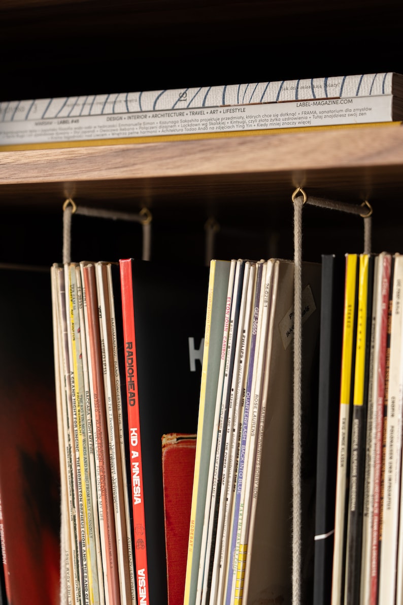 May include: A wooden shelf displays a collection of vinyl records and magazines. The records are in various colors, with titles like "Radiohead" and "Kid A Mnesia" visible. A magazine with the text "LABEL-MAGAZINE.COM" is on top.