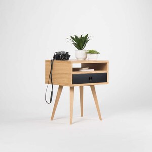 Puede incluir: Una mesita de noche de madera con un caj&oacute;n negro y un estante. La mesita de noche tiene cuatro patas y est&aacute; decorada con dos plantas en maceta y una c&aacute;mara.
