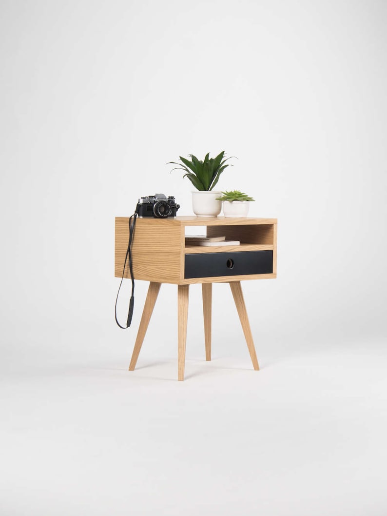 Puede incluir: Una mesita de noche de madera con un caj&oacute;n negro y un estante. La mesita de noche tiene cuatro patas y est&aacute; decorada con dos plantas en maceta y una c&aacute;mara.