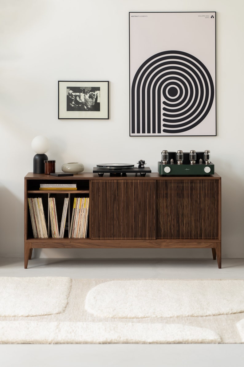 May include: A mid-century modern style record player console with a dark wood finish. The console features a built-in record player, a green amplifier, and storage for vinyl records. A black and white art print hangs on the wall.