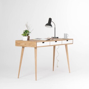 Puede incluir: Un escritorio de madera clara con tres cajones blancos y patas c&oacute;nicas. Una l&aacute;mpara negra con pantalla blanca se encuentra sobre el escritorio, junto con una taza blanca y un libro.