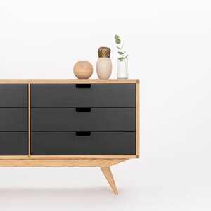 May include: A six-drawer dresser with a light wood frame and black drawers. The dresser has a modern design with angled legs. There are three decorative items on top of the dresser, including a small vase with a sprig of greenery, a ceramic vase, and a round wooden bowl.