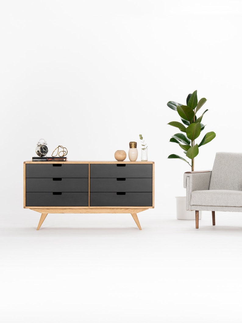 May include: A six-drawer dresser with a light wood frame and dark gray drawers. The dresser has a modern design with tapered legs. The dresser is sitting on a white floor in front of a white wall.